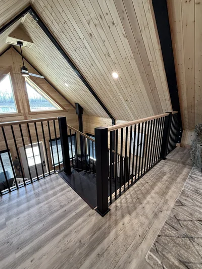 Finished timber loft stair landing with black railings, natural wood handrails, vaulted wood ceiling, and triangular windows